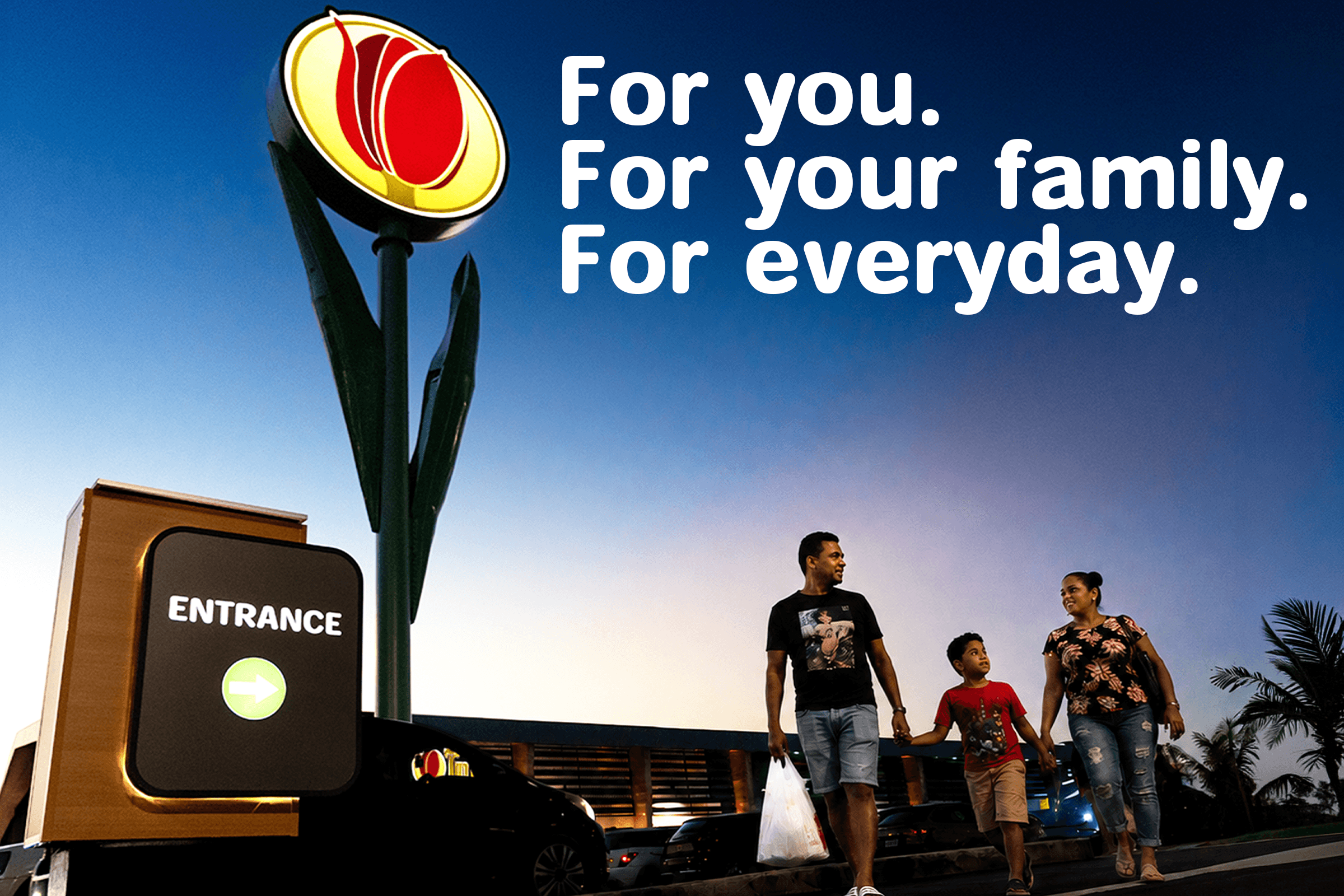 Illuminated Tulip sign at the store entrance as a family walks by at dusk, with the words: For you. For your family. For everyday.